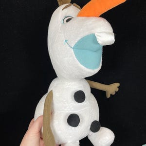 May include: A plush Olaf doll from the movie Frozen. The snowman has a white body, an orange carrot nose, and a blue mouth. He has three black buttons and brown twig arms and hair. The doll is held by a hand.
