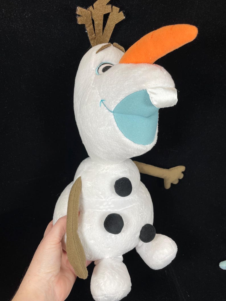 May include: A plush Olaf doll from the movie Frozen. The snowman has a white body, an orange carrot nose, and a blue mouth. He has three black buttons and brown twig arms and hair. The doll is held by a hand.