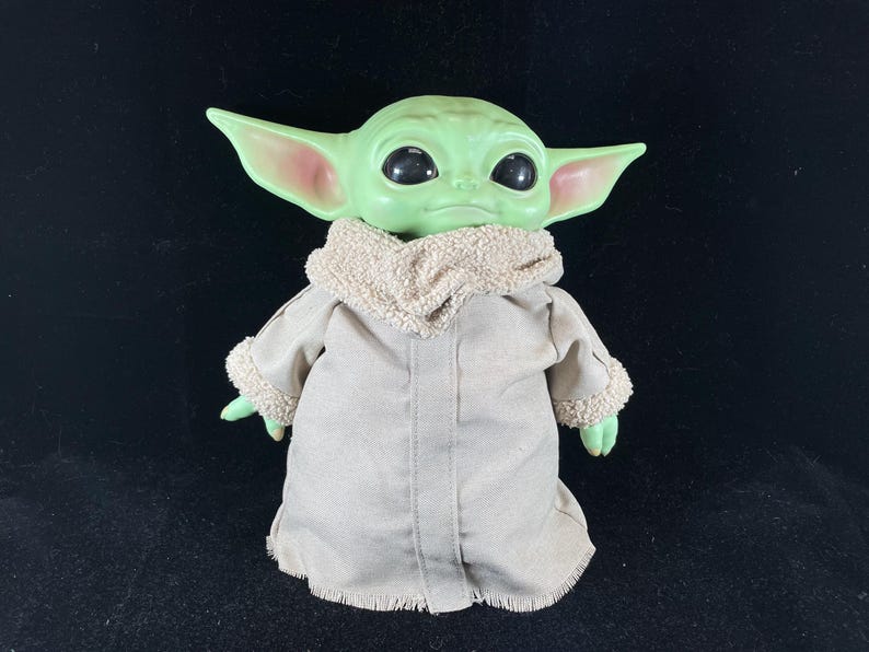May include: A green-skinned doll with large black eyes and pointed ears, dressed in a tan coat with a fluffy collar and cuffs. The doll is a character from a popular science fiction franchise.