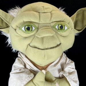 Op de afbeelding: Een Yoda-knuffelpop uit Star Wars. De pop heeft een lichtgroen gezicht, grote oren en wit haar. Hij draagt een beige gewaad en heeft geborduurde groene ogen. De handen van de pop zijn gevouwen.