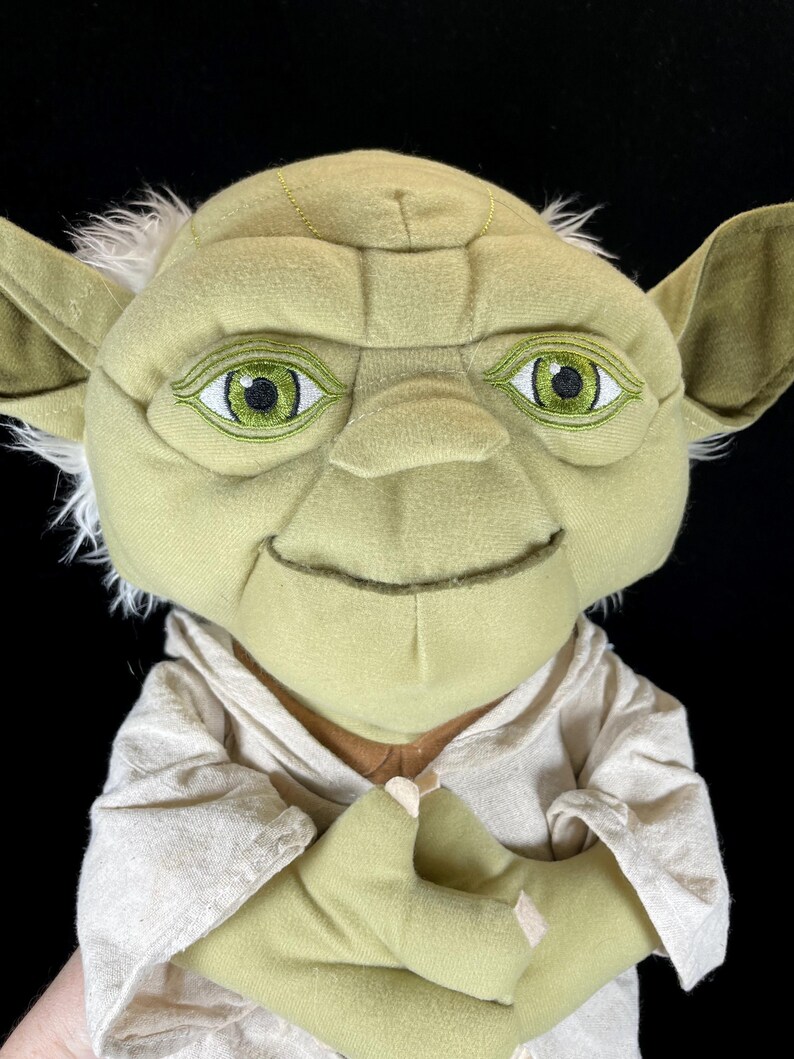 Op de afbeelding: Een Yoda-knuffelpop uit Star Wars. De pop heeft een lichtgroen gezicht, grote oren en wit haar. Hij draagt een beige gewaad en heeft geborduurde groene ogen. De handen van de pop zijn gevouwen.