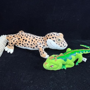 May include: Two plush reptile toys. One is a tan and brown spotted gecko with a long, cream-colored tail. The other is a green and blue lizard with a detailed design. Both are lying on a black surface.