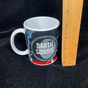 Op de afbeelding: Witte keramische mok met een zwarte achtergrond met de tekst "Darth Sidious" in een witte ovaal. De mok heeft een wit handvat en een rood en blauw bliksemontwerp. De mok is ongeveer 7,6 cm hoog.
