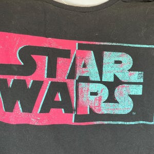 Op de afbeelding: Zwart t-shirt met het "STAR WARS" logo. Het logo is in twee&euml;n gedeeld, de linkerkant roze, de rechterkant turquoise, met een distressed, getextureerd effect.