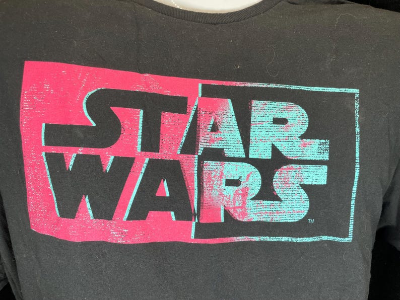 Op de afbeelding: Zwart t-shirt met het "STAR WARS" logo. Het logo is in twee&euml;n gedeeld, de linkerkant roze, de rechterkant turquoise, met een distressed, getextureerd effect.