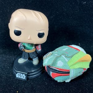 May include: A Star Wars themed collectible set featuring a Funko Pop figure and a plush helmet. The Funko Pop figure is of a bald character in a green and red outfit. The plush helmet is green, red, and orange.