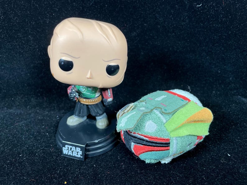 May include: A Star Wars themed collectible set featuring a Funko Pop figure and a plush helmet. The Funko Pop figure is of a bald character in a green and red outfit. The plush helmet is green, red, and orange.