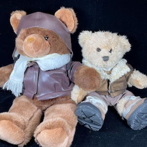 Puede incluir: Dos osos de peluche. Uno es marrón con un sombrero, chaqueta y bufanda de aviador de cuero marrón. El otro es de color canela con una chaqueta, pantalones y botas marrones. Ambos osos tienen ojos y nariz negros.