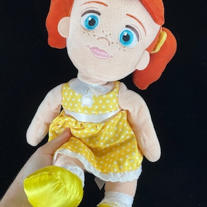 May include: A plush doll with red hair styled in pigtails, blue eyes, and freckles. The doll wears a yellow dress with white polka dots, a white collar, and yellow shoes. The doll is a character from a popular animated film.