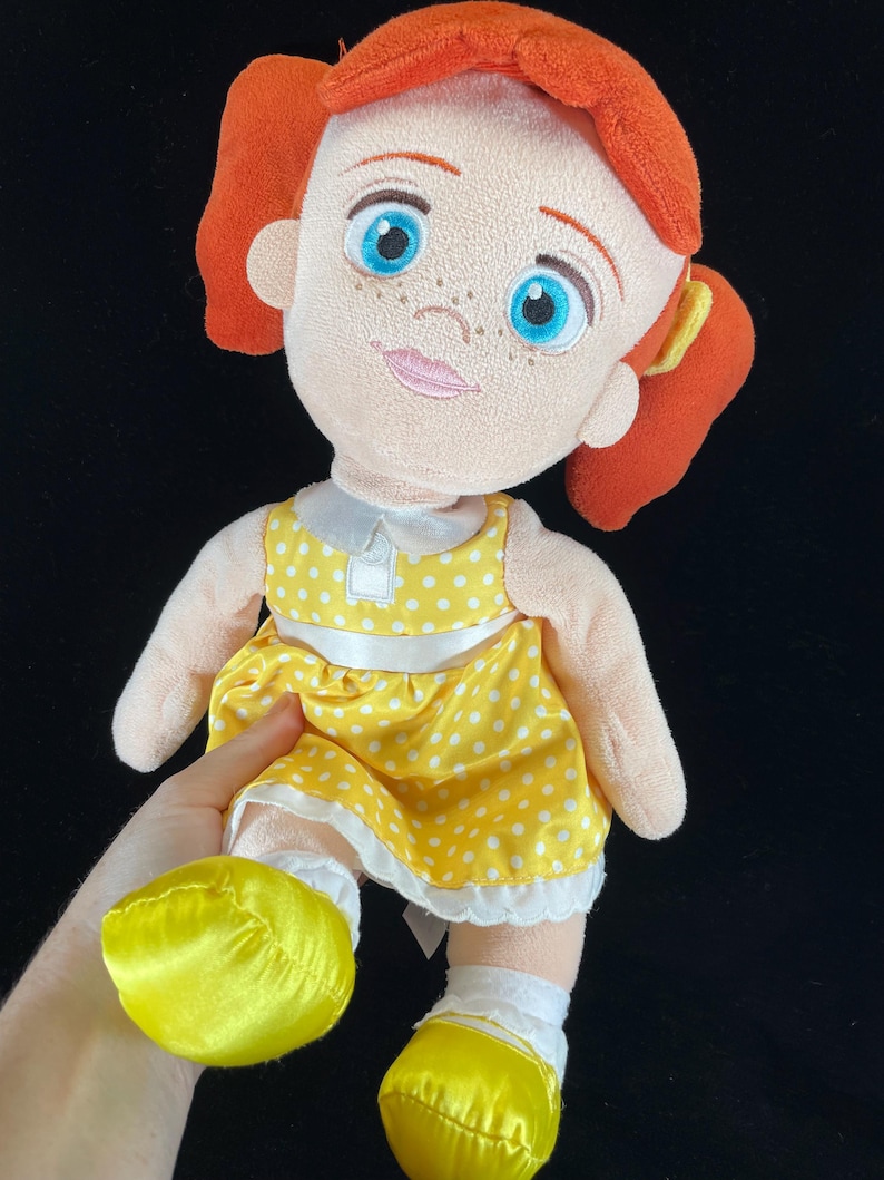 May include: A plush doll with red hair styled in pigtails, blue eyes, and freckles. The doll wears a yellow dress with white polka dots, a white collar, and yellow shoes. The doll is a character from a popular animated film.