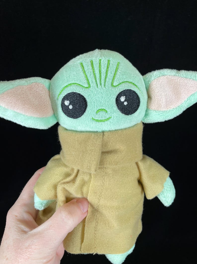 Op de afbeelding: Een pluche pop van een personage met grote zwarte ogen, grote oren en een lichtgroene teint. De pop draagt een bruin jasje. De pop wordt door een persoon vastgehouden.