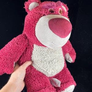 May include: A plush, pink teddy bear with a white belly and a dark pink nose. The bear has a friendly expression and is made of soft, fuzzy material. The bear's ears and feet are a lighter shade of pink.