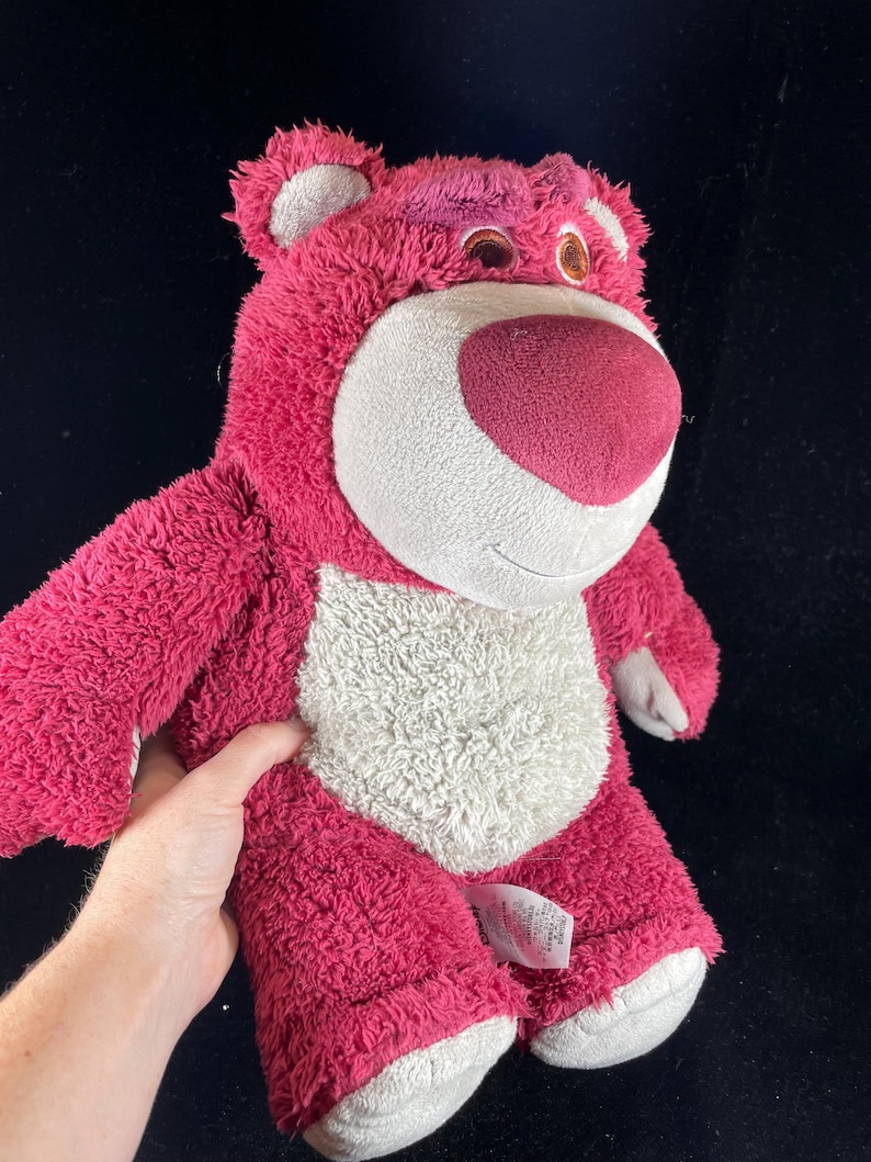 May include: A plush, pink teddy bear with a white belly and a dark pink nose. The bear has a friendly expression and is made of soft, fuzzy material. The bear's ears and feet are a lighter shade of pink.