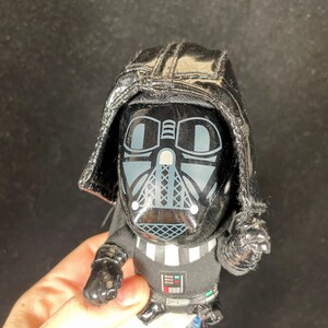 Op de afbeelding: Een kleine, zwarte Darth Vader-figuur met een glanzende helm en masker. De figuur heeft een zwarte cape en een gedetailleerde borstplaat met een zilver en wit ontwerp. Verzamelobject.