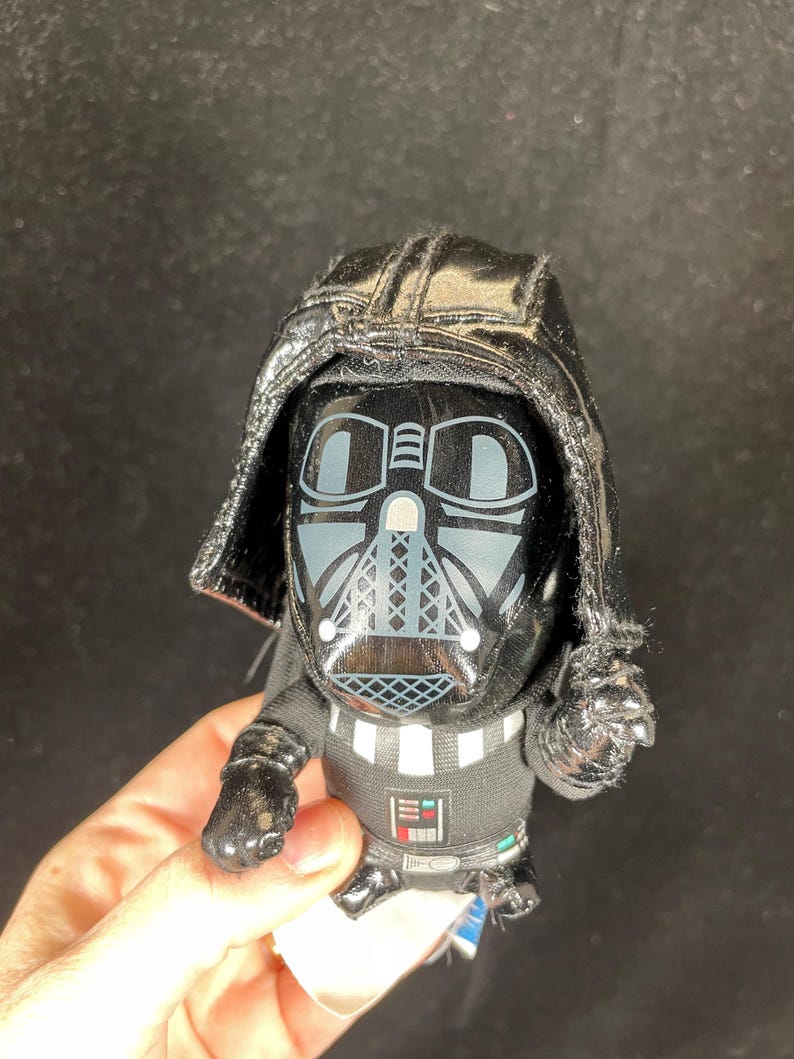 Op de afbeelding: Een kleine, zwarte Darth Vader-figuur met een glanzende helm en masker. De figuur heeft een zwarte cape en een gedetailleerde borstplaat met een zilver en wit ontwerp. Verzamelobject.
