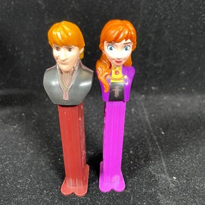 May include: Two PEZ dispensers featuring characters from the animated film "Frozen." One dispenser has a red base and depicts a male character with orange hair. The other has a purple base and features a female character with red hair.