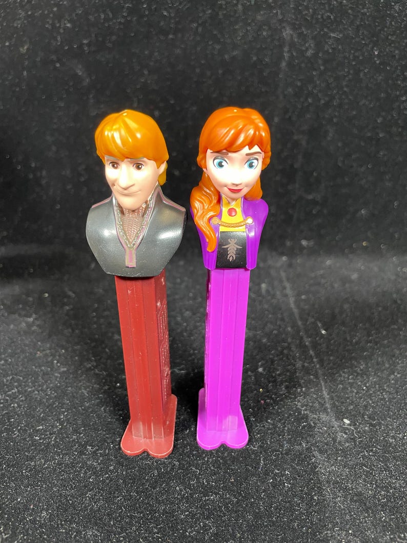 May include: Two PEZ dispensers featuring characters from the animated film "Frozen." One dispenser has a red base and depicts a male character with orange hair. The other has a purple base and features a female character with red hair.