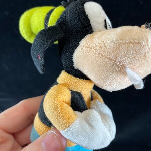 Puede incluir: Un peluche de Goofy, un personaje de dibujos animados de Disney. El juguete tiene una cabeza negra con una cara blanca, un sombrero amarillo y una camisa a rayas amarillas y negras. El juguete lleva pantalones azules y guantes blancos.