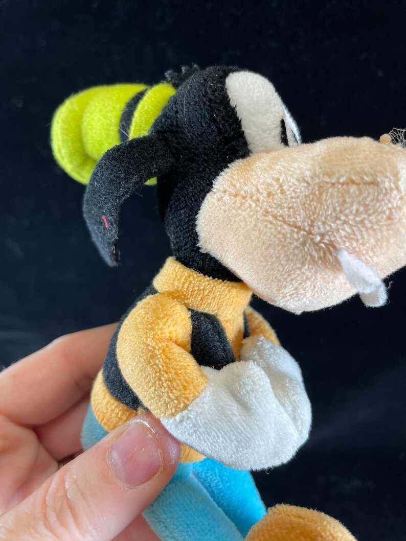 Puede incluir: Un peluche de Goofy, un personaje de dibujos animados de Disney. El juguete tiene una cabeza negra con una cara blanca, un sombrero amarillo y una camisa a rayas amarillas y negras. El juguete lleva pantalones azules y guantes blancos.