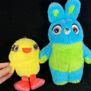 May include: Two plush toys: a yellow chick with orange beak and red feet, and a blue and green bunny with large eyes. The chick is held by a hand. The toys are on a black background.