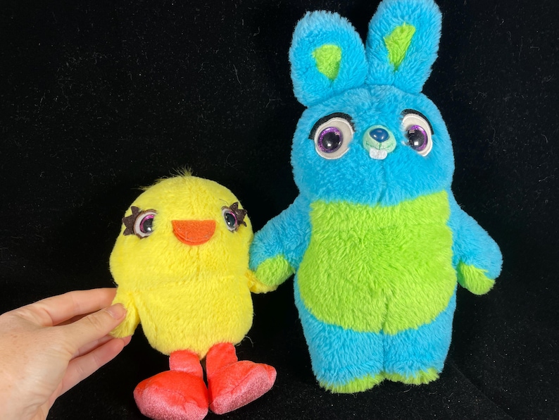 May include: Two plush toys: a yellow chick with orange beak and red feet, and a blue and green bunny with large eyes. The chick is held by a hand. The toys are on a black background.