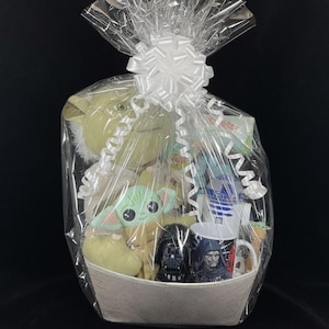 Op de afbeelding: Een cadeaupakket gevuld met Star Wars-thema merchandise. De mand bevat pluche speelgoed van Yoda en Grogu, mokken met Darth Vader, R2-D2 en andere personages, en andere diverse items. De mand is verpakt in doorzichtig cellofaan en vastgebonden met een witte strik.