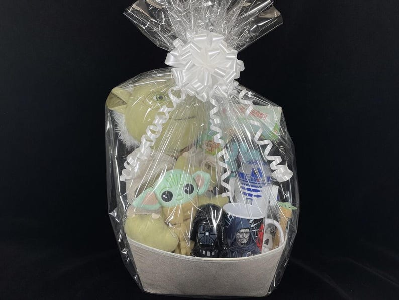 Op de afbeelding: Een cadeaupakket gevuld met Star Wars-thema merchandise. De mand bevat pluche speelgoed van Yoda en Grogu, mokken met Darth Vader, R2-D2 en andere personages, en andere diverse items. De mand is verpakt in doorzichtig cellofaan en vastgebonden met een witte strik.