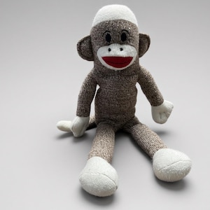 May include: A stuffed monkey toy crafted from brown heathered knit fabric. It features a white knit cap, white hands, and white feet. The face has a white oval muzzle, a red stitched mouth, and two black circular eyes set above the muzzle.