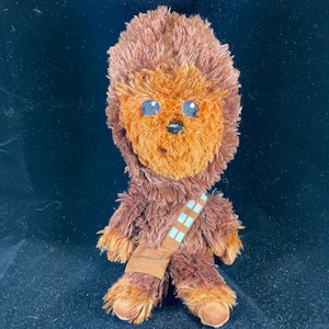 Könnte beinhalten: Ein braunes Plüsch-Chewbacca-Spielzeug mit blauen Augen und einem braunen Bandolero. Das Spielzeug sitzt auf einer schwarzen Oberfläche.