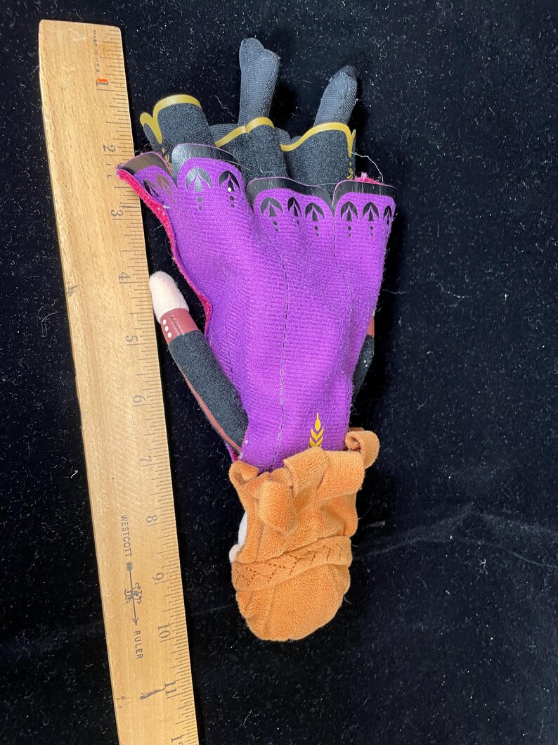 May include: A purple and black glove with gold trim, featuring a brown cuff and a decorative pattern. The glove is approximately 5 inches long, with a wooden ruler in the image for scale.