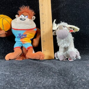 Puede incluir: Dos peluches: un mono marr&oacute;n con una camiseta de Tune Squad y un bal&oacute;n de baloncesto, y un reno gris. El mono mide aproximadamente 13 cm de alto. El reno mide aproximadamente 8 cm de alto. Ambos juguetes est&aacute;n sobre una superficie negra.