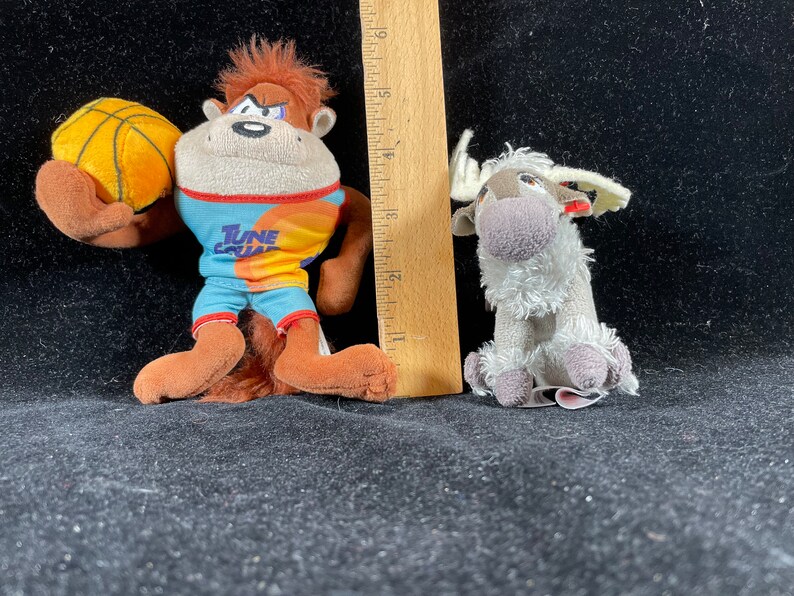 Puede incluir: Dos peluches: un mono marr&oacute;n con una camiseta de Tune Squad y un bal&oacute;n de baloncesto, y un reno gris. El mono mide aproximadamente 13 cm de alto. El reno mide aproximadamente 8 cm de alto. Ambos juguetes est&aacute;n sobre una superficie negra.