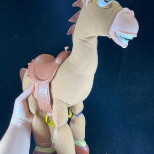 May include: A plush toy horse with a brown body, saddle, and mane. The horse has a light pink nose, white teeth, and blue eyes. The saddle has a cactus design. The toy is approximately 12 inches tall.
