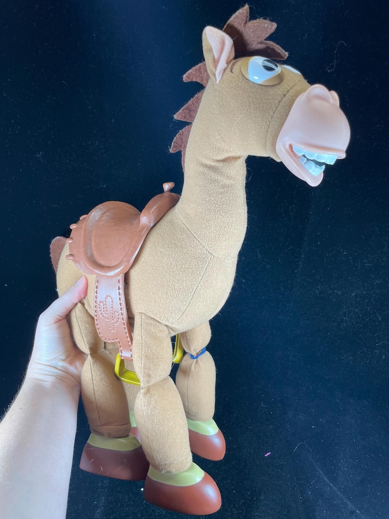 May include: A plush toy horse with a brown body, saddle, and mane. The horse has a light pink nose, white teeth, and blue eyes. The saddle has a cactus design. The toy is approximately 12 inches tall.