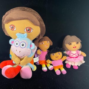 May include: Four plush toys of Dora the Explorer and her friends. The largest toy is Dora, holding a purple and blue plush monkey. The other three toys are smaller and depict Dora's friends, all wearing pink and purple dresses.
