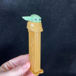 Op de afbeelding: Een goudkleurige PEZ-dispenser met een groene Grogu-karakterkop. De dispenser heeft een rechthoekige behuizing met reli&euml;fdetails en een drielobbige basis. De Grogu-kop is lichtgroen met grote ogen.