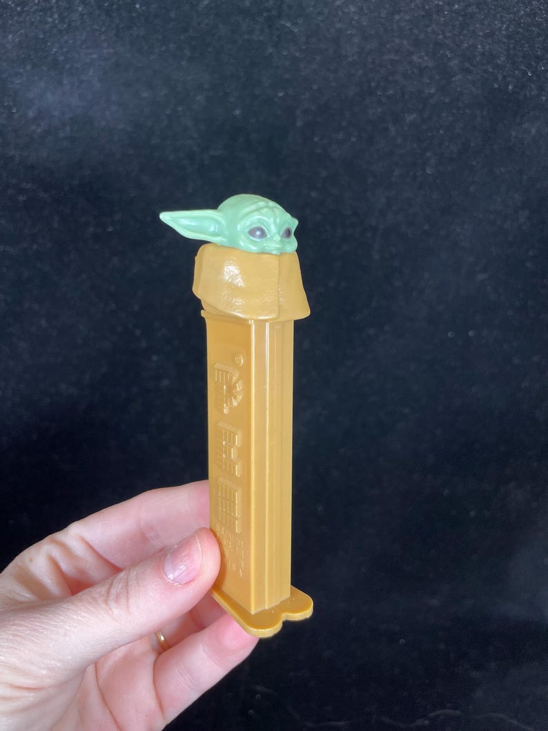 Op de afbeelding: Een goudkleurige PEZ-dispenser met een groene Grogu-karakterkop. De dispenser heeft een rechthoekige behuizing met reli&euml;fdetails en een drielobbige basis. De Grogu-kop is lichtgroen met grote ogen.