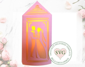 May include: A pink papercraft wedding lantern with a couple silhouette inside. The lantern has a yellow glow and is decorated with a white crown on top. The text "Digitally Delivered Template SVG Bella Carta Design" is on a green circle.