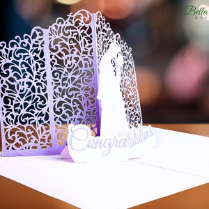Left side view of a lavender and white pop-up card, featuring a detailed silhouette of a bride and groom. The card is closed, showcasing intricate, ornate cut-out designs on the front and side.