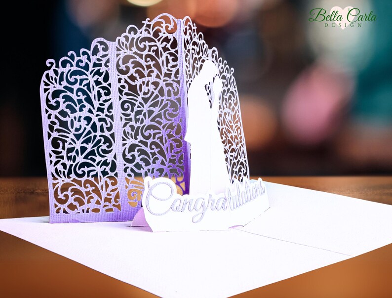 Left side view of a lavender and white pop-up card, featuring a detailed silhouette of a bride and groom. The card is closed, showcasing intricate, ornate cut-out designs on the front and side.