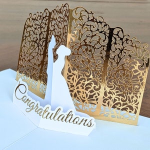 Right side view of an elegant gold and white pop-up card featuring a detailed silhouette of a bride and groom. The front reads 'Congratulations' in gold script.