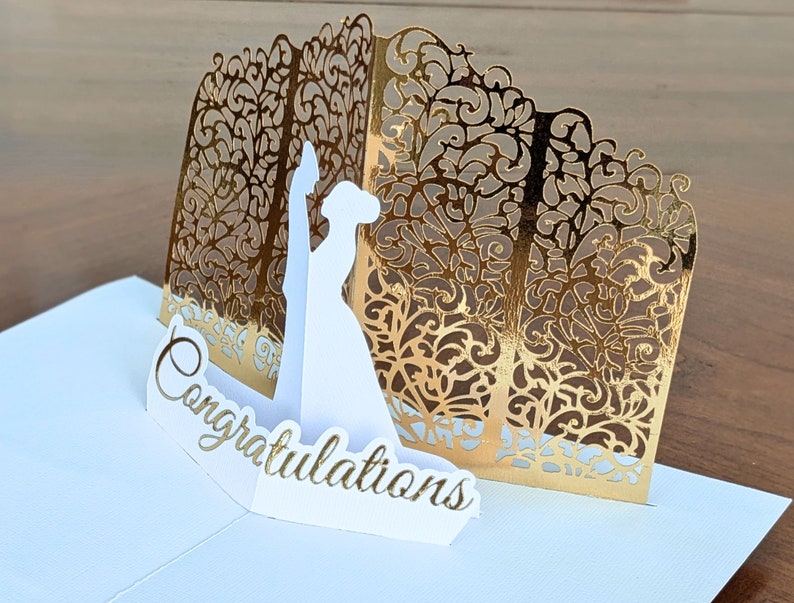 Right side view of an elegant gold and white pop-up card featuring a detailed silhouette of a bride and groom. The front reads 'Congratulations' in gold script.