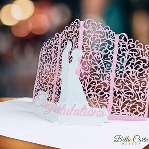 May include: A pop-up greeting card featuring a white silhouette of a couple standing before a light pink, laser-cut, intricate floral archway. The word Congratulations is printed in pink script on a white base at the front of the card.