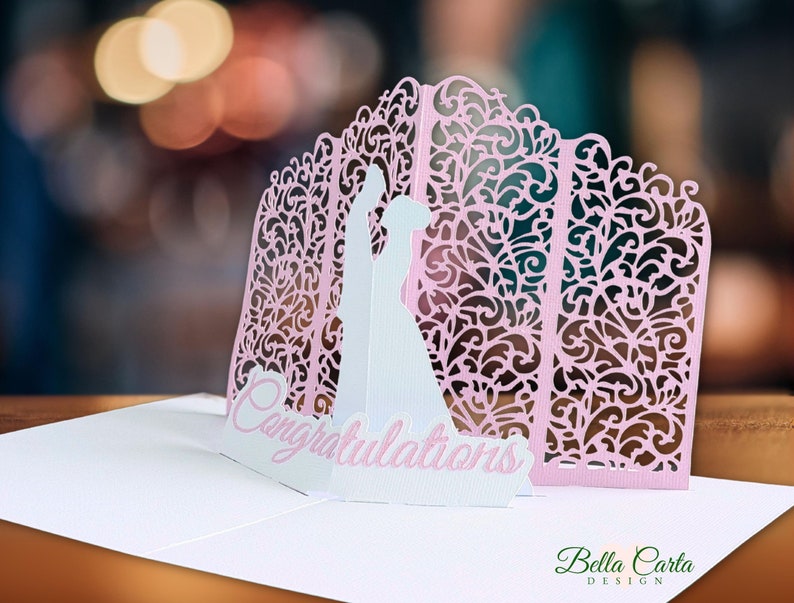 May include: A pop-up greeting card featuring a white silhouette of a couple standing before a light pink, laser-cut, intricate floral archway. The word Congratulations is printed in pink script on a white base at the front of the card.