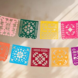 May include: A colorful paper banner with cut-out designs, featuring the phrase "Dia de Muertos" in orange and purple. The banner is made of pink, green, yellow, red, blue, and orange paper.
