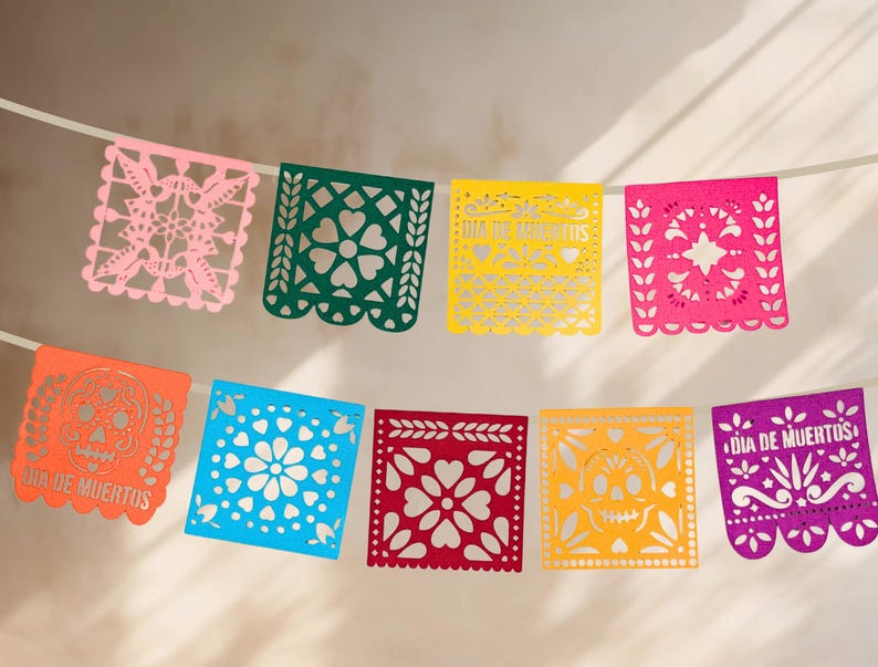 May include: A colorful paper banner with cut-out designs, featuring the phrase "Dia de Muertos" in orange and purple. The banner is made of pink, green, yellow, red, blue, and orange paper.