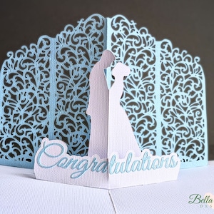 May include: A light blue pop-up greeting card featuring a laser-cut intricate filigree background and a central silhouette of two figures facing each other. The word Congratulations is printed in a script font on a white base in the foreground.
