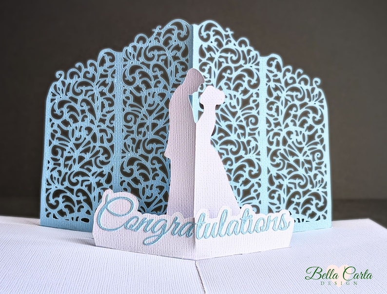 May include: A light blue pop-up greeting card featuring a laser-cut intricate filigree background and a central silhouette of two figures facing each other. The word Congratulations is printed in a script font on a white base in the foreground.