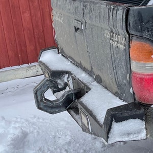 Puede incluir: Una camioneta Toyota Tacoma PreRunner negra con un parachoques trasero cubierto de nieve y un enganche de remolque de alta resistencia. La camioneta está estacionada frente a un granero de madera rojo.