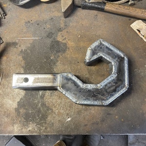 Puede incluir: Un gancho de metal con un mango soldado. El gancho está hecho de acero y tiene un aspecto áspero e industrial. El gancho está sobre una mesa de trabajo de metal.